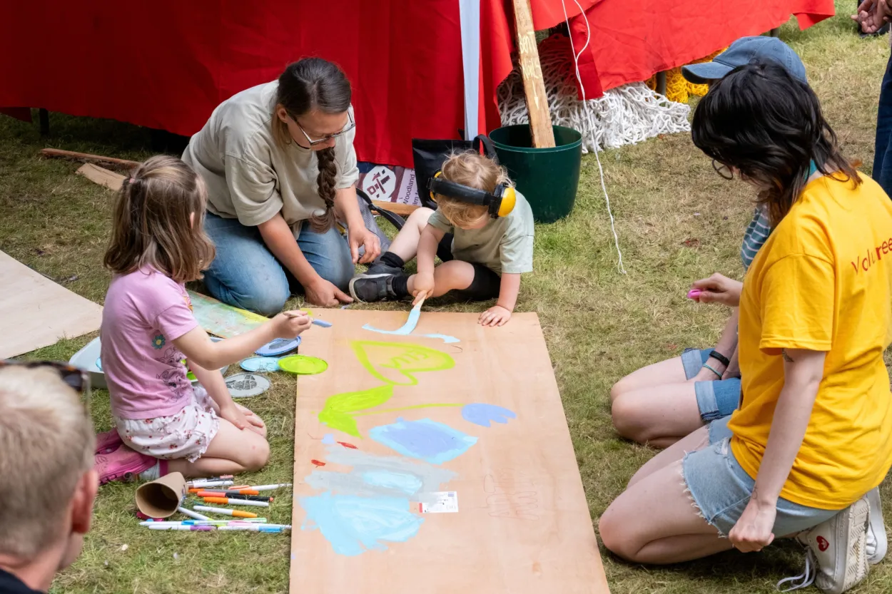 An adult and two children are drawing on a large piece of wood, outside on some grass.