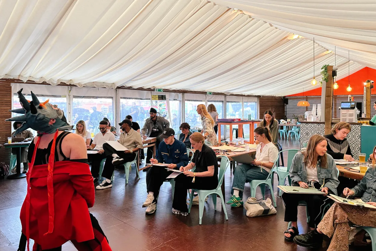 A model is Posing in a Bright Red jacket wearing a sculptural headset. Multiple people are sitting drawing them.