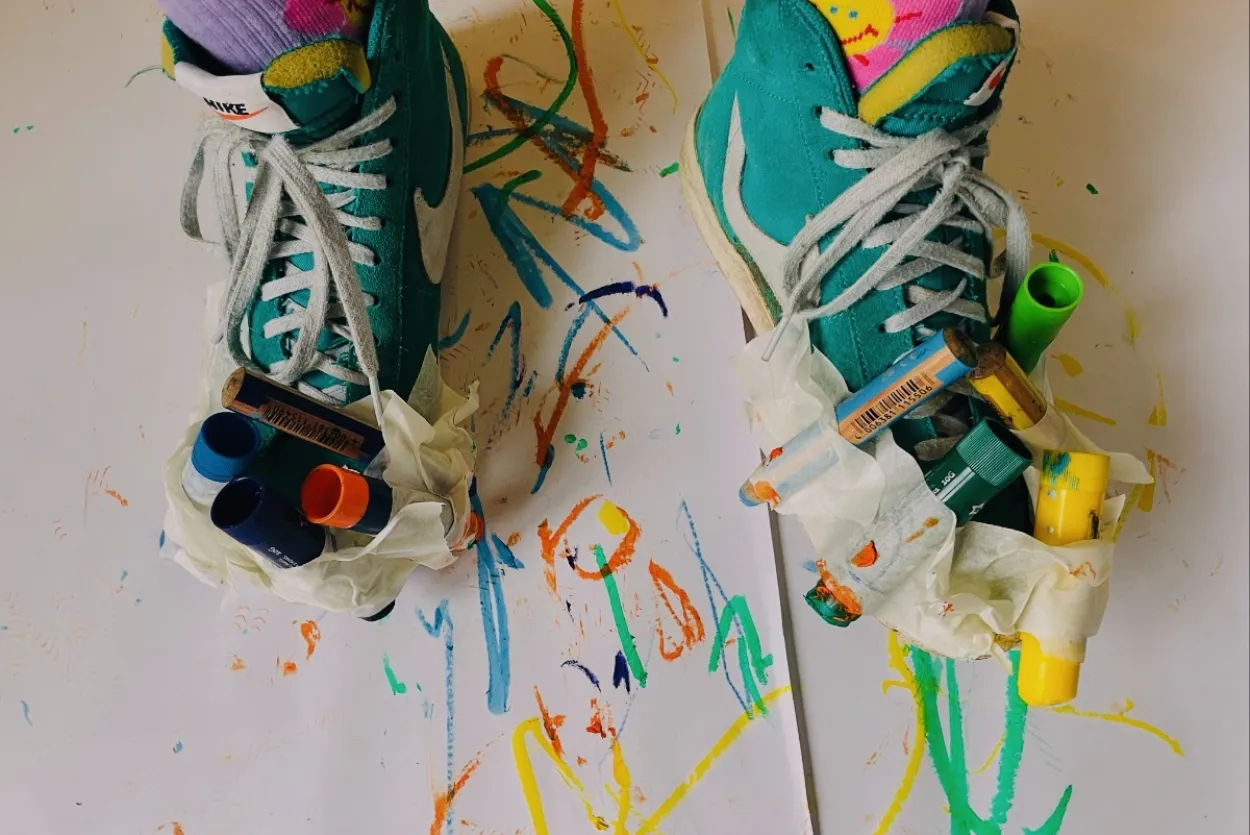 photo of a pair of feet from above- wearing pink and purple socks and green trainers. the trainers have pens and crayons taped around the edge of them and there is paper underneath covered in colourful marks from the attached pens.