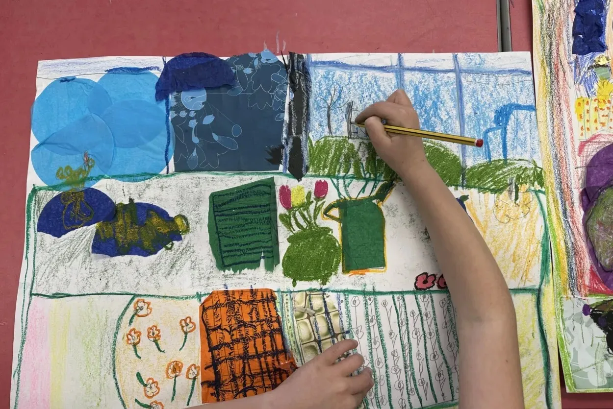 A child's hand holding a pencil is drawing on a colourful, large piece of paper filled with various shapes and textures, including plants and abstract designs.