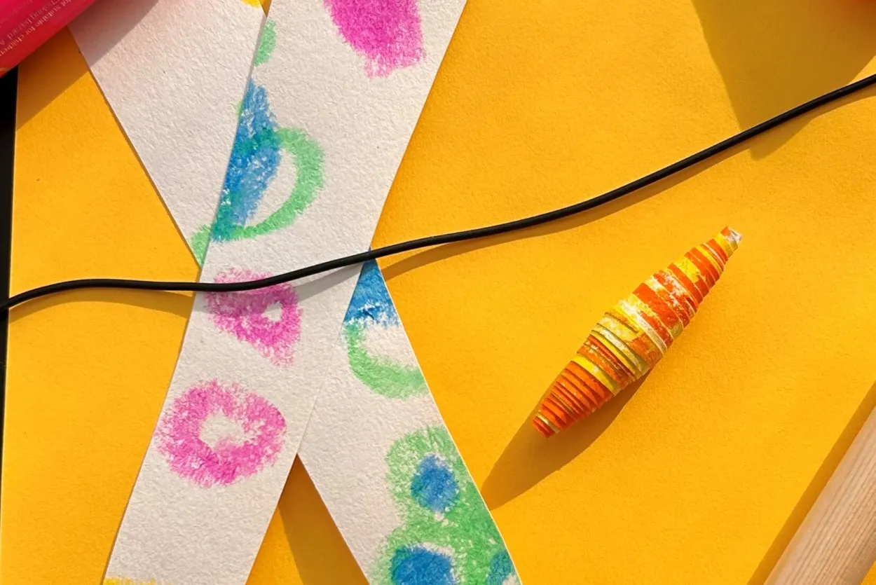Strips of paper with colourful patterns next to a pen, glue stick, paper bead, cord and a jewellery making tool.