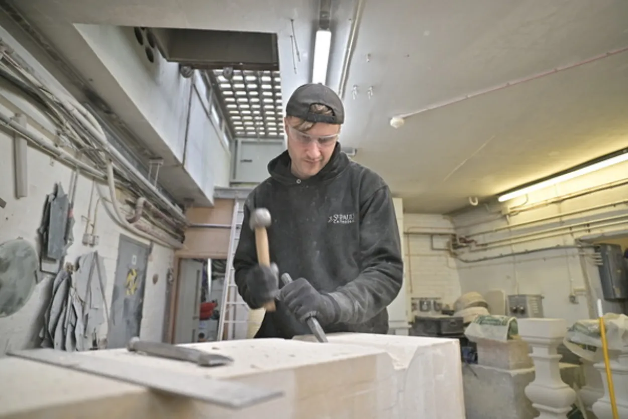 White male in mid 20s, wearing a black back to front baseball cap, hoodie and and gloves, using a hammer and chisel to carve into a block of white stone