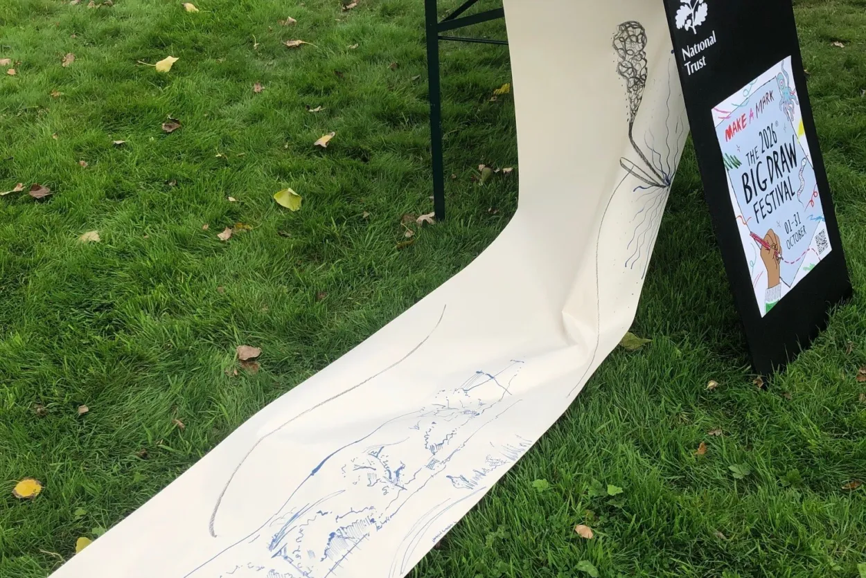 A large roll of paper with ink marks and drawing, hanging off a table with a poster for The Big Draw 2026 beside it.