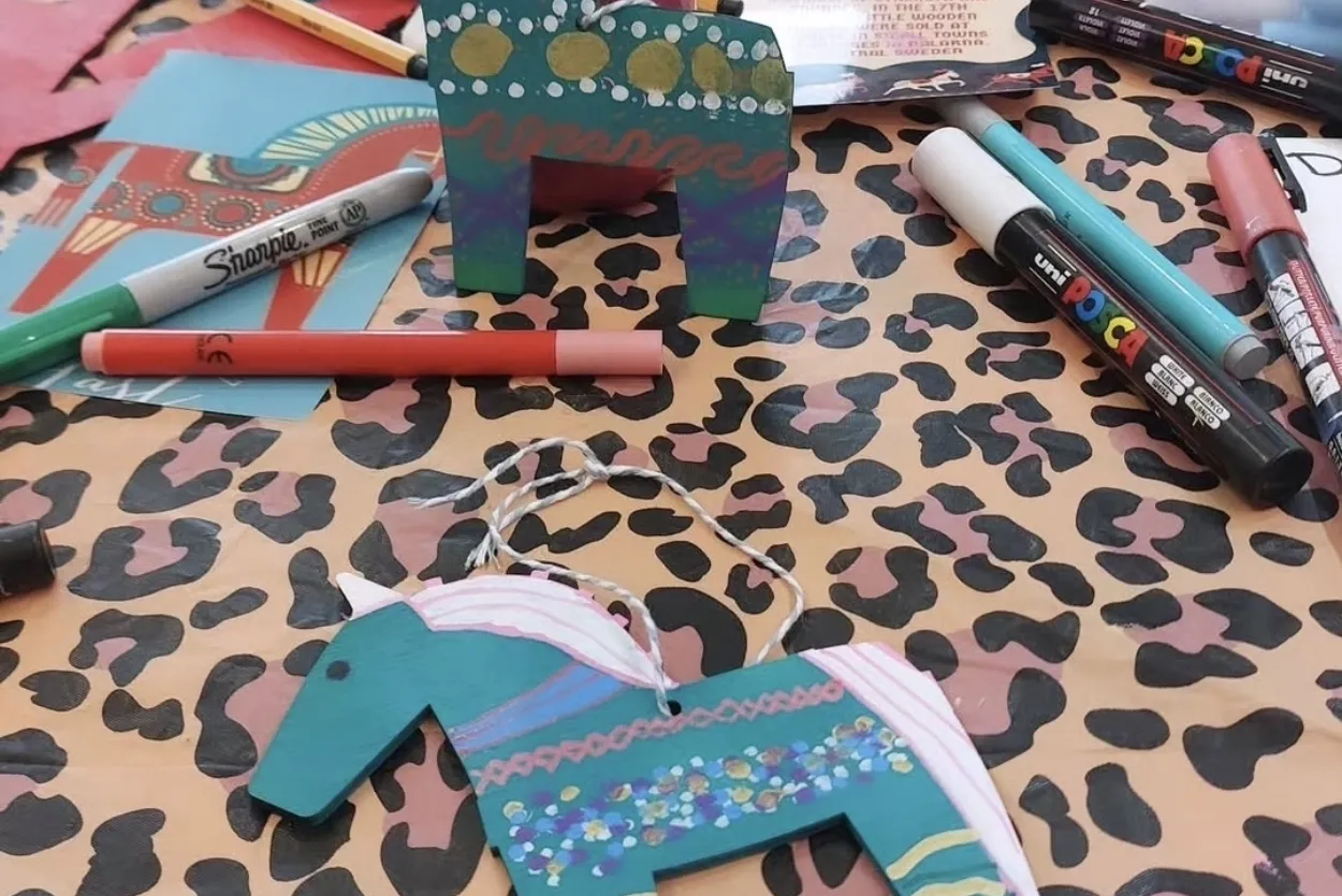 Two hand drawn dala horses on a leopard print tablecloth surrounded by pens.