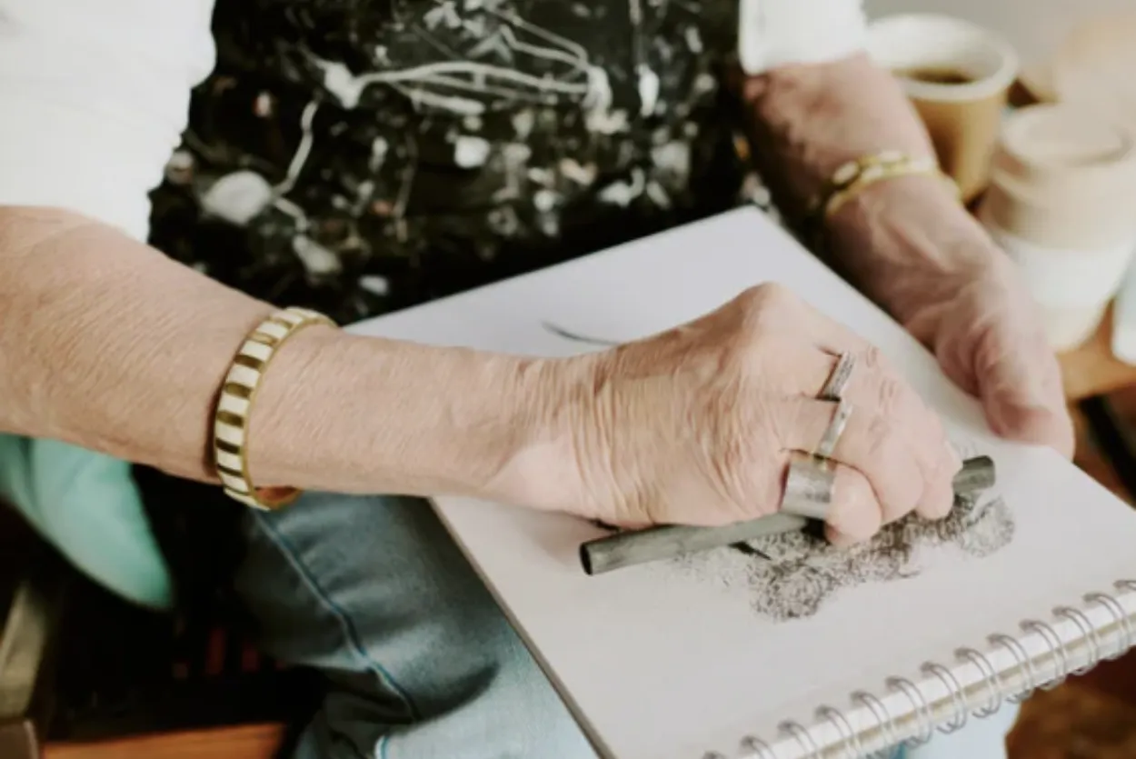 A close up shot of an adult holding a piece of charcoal and smudging it on a sketchbook. The person is wearing a white long-sleeved shirt, blue jeans, a paint-splattered black apron, and several rings and bracelets.