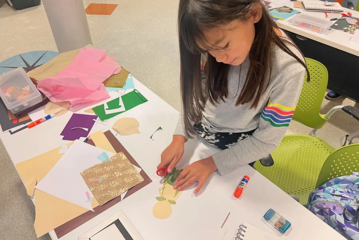 A little girl works with scissors and green paper as she creates her paper doll on a table. There are different colored sheets of paper with a variety of textures next to her hands.