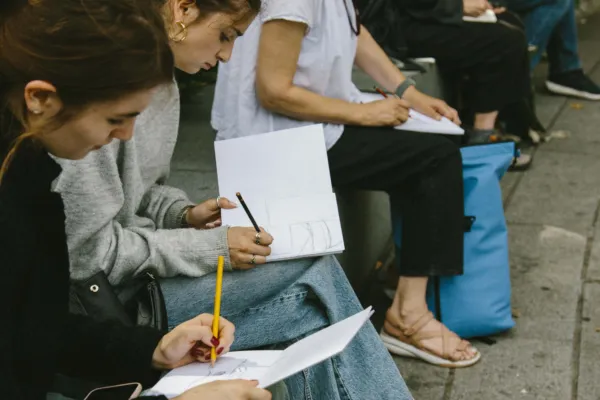 Adults sat on a bench during a sketch crawl with sketchbooks in hand focusing and drawing something on the opposite street out of view.