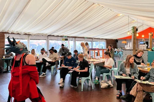 A model is Posing in a Bright Red jacket wearing a sculptural headset. Multiple people are sitting drawing them.