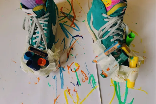 photo of a pair of feet from above- wearing pink and purple socks and green trainers. the trainers have pens and crayons taped around the edge of them and there is paper underneath covered in colourful marks from the attached pens.