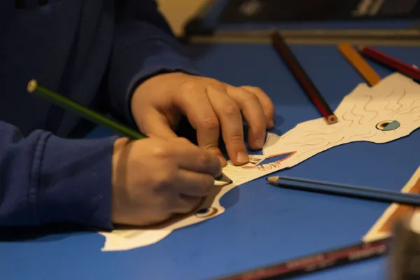 a child's hands draws on a dragon mask with various coloured pencils scattered around