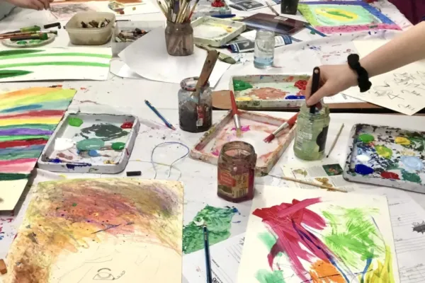 A messy art table with colourful paintings, splatters of paint, brushes, jars of paint, and various art supplies scattered around.