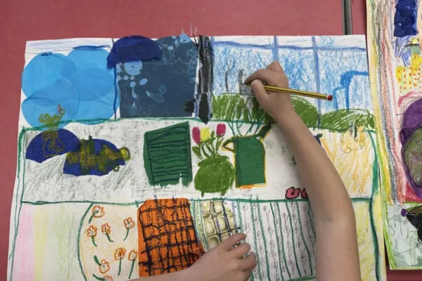 A child's hand holding a pencil is drawing on a colourful, large piece of paper filled with various shapes and textures, including plants and abstract designs.