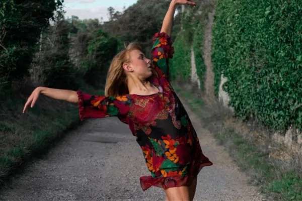 An image of a young woman in a short flowing red long-sleeved dress. She is on a country lane with greenery behind her and is striking a contemporary dance pose with her arms spread above and body leaning back.