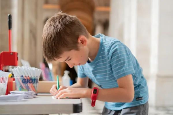 Young boy wearing a blue top, bent over a table drawing using pencils and paper.