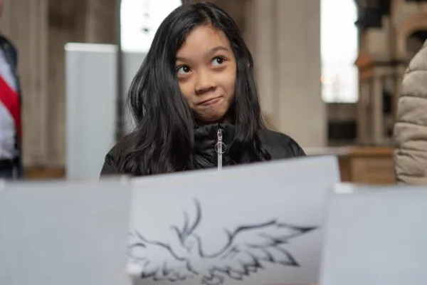 Young girl wearing a black zip up jacket, holding in front of her body a piece of drawing paper and looking thoughtfully out of shot. The Background is the nave of St Paul's Cathedral