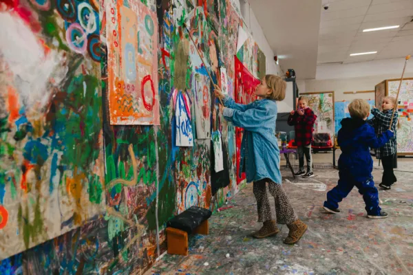 Children engaged in painting a large, colourful design on a wall, surrounded by art supplies in a cheerful studio setting.