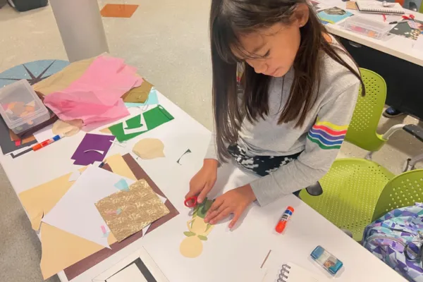 A little girl works with scissors and green paper as she creates her paper doll on a table. There are different colored sheets of paper with a variety of textures next to her hands.